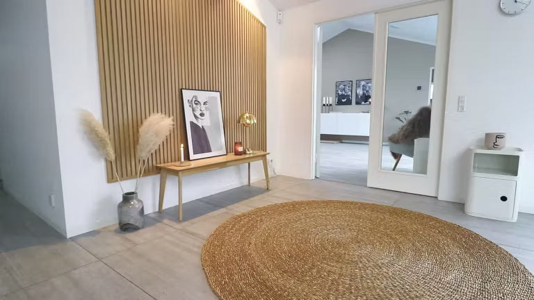 Minimal Nordic living space with a round jute rug, wooden bench holding pampas grass, framed art, and a slatted Akupanel acoustic wood accent wall.